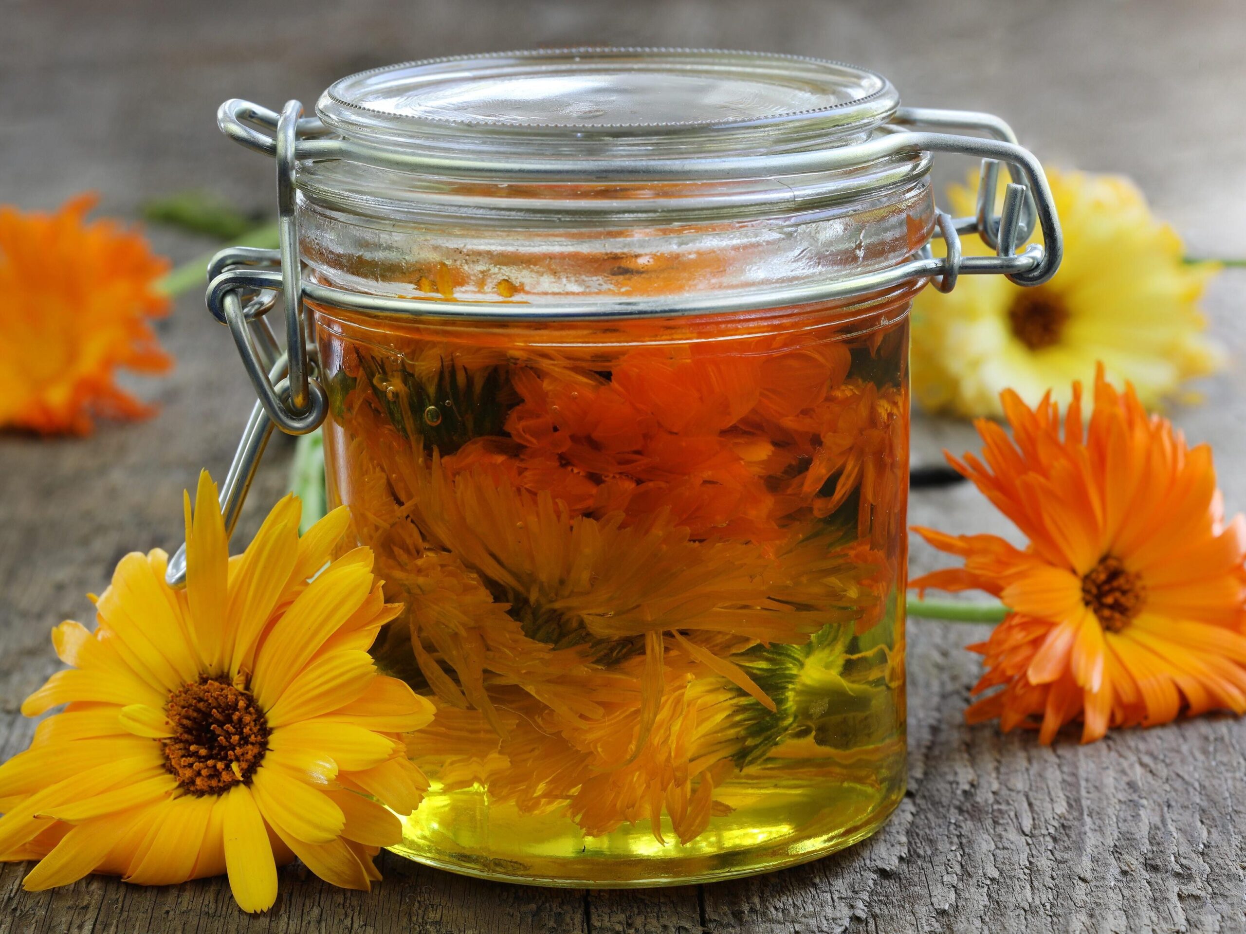 Calendula flowers and golden herbal infused oil in glass jars on a rustic wooden table.
